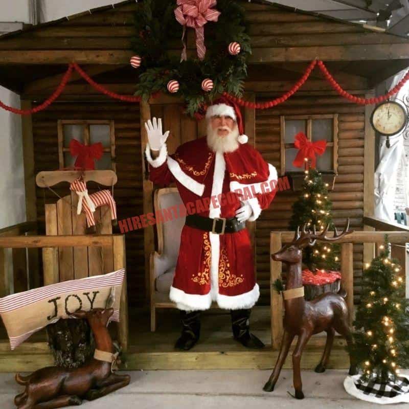 Real beard Santa for hire in Cape Coral, FL waving from a log cabin porch beside a wooden reindeer.