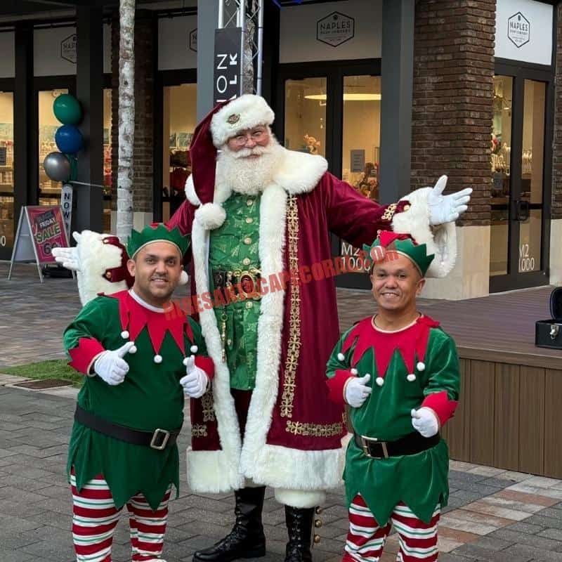 Santa Claus impersonator for hire in Cape Coral, FL in a burgundy coat, thumbs up outdoors with two elves in green.