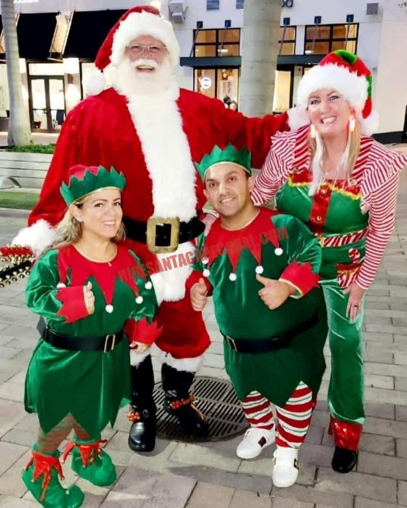 Santa for hire in Lehigh Acres, FL stands with three elves on a plaza at dusk.